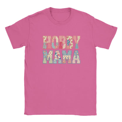 A pink short-sleeve crewneck t-shirt featuring a colorful graphic text print that reads 'HOBBY MAMA' in a patchwork pattern of various small colored squares and floral motifs.