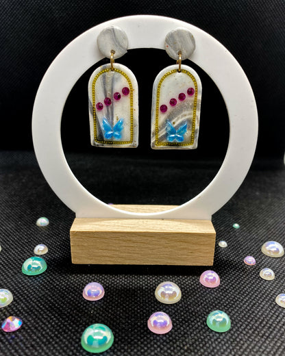 Close-up of white clay arch earring with blue butterfly, pink gems, silver shimmer and gold-tone outline