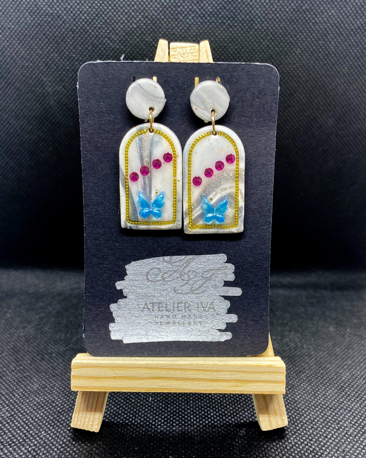 Pair of white clay arch earrings with blue butterflies, pink gems and gold-tone outlines on display card