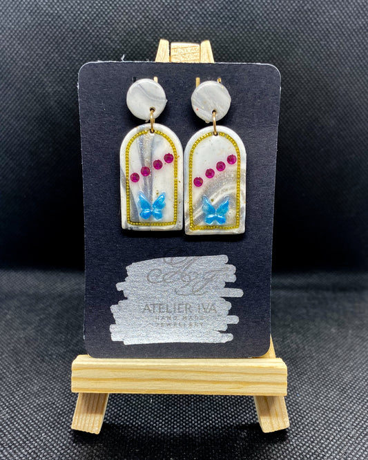 Pair of white clay arch earrings with blue butterflies, pink gems and gold-tone outlines on display card