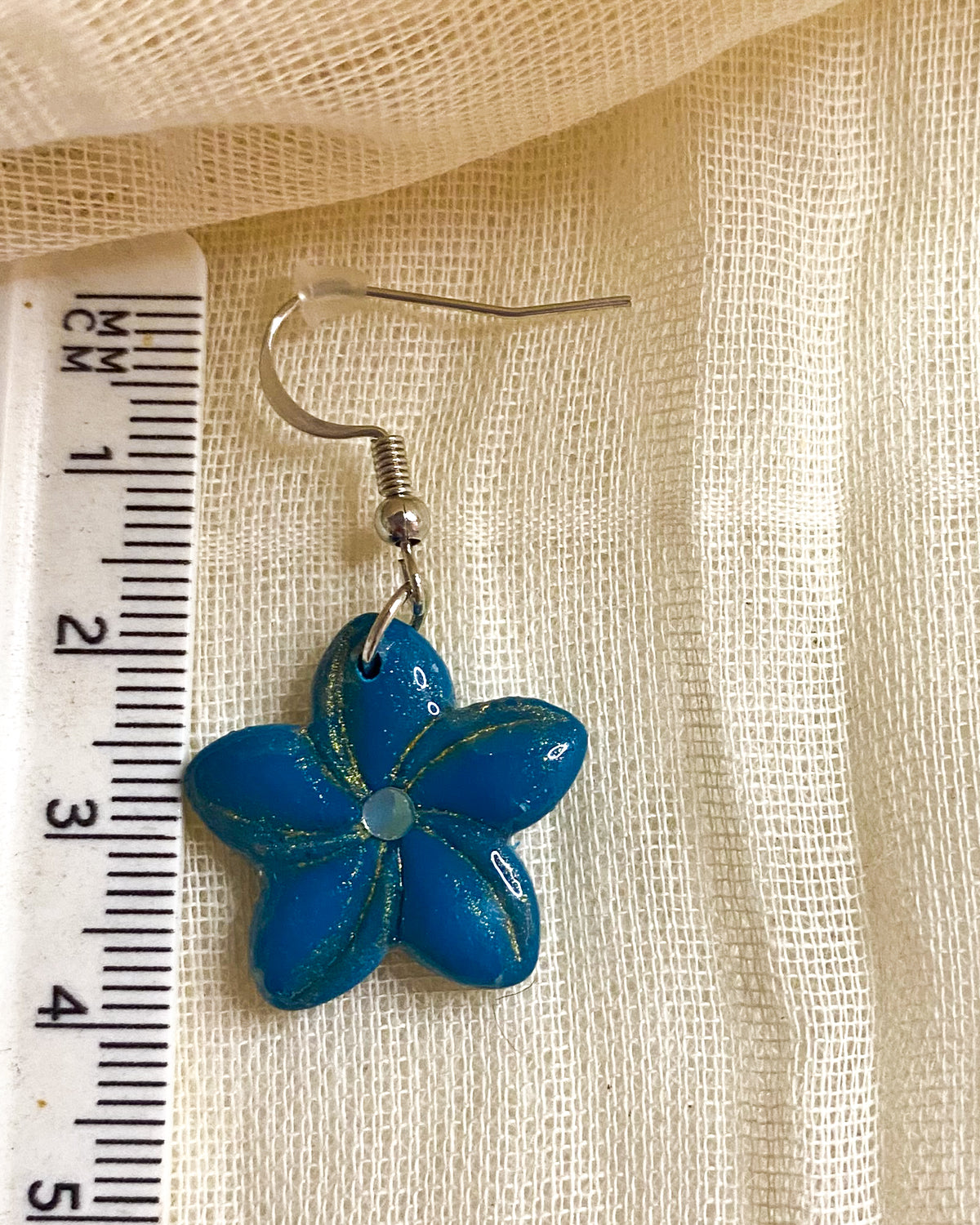 Close-up of turquoise five-petal clay flower earring with glitter-lined details, pale centre and silver-tone hook beside a ruler