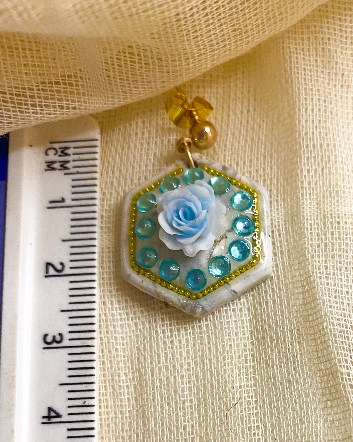 Close-up of white marbled hexagon clay earring with pale blue rose, aqua gems and gold-tone beaded border beside a ruler
