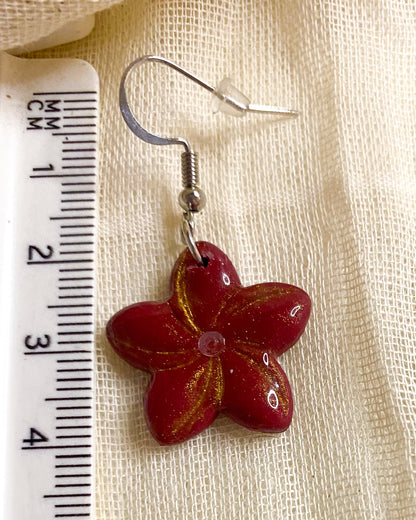 Close-up of deep red five-petal clay flower earring with gold dust detailing, rhinestone centre and silver-tone hook beside a ruler