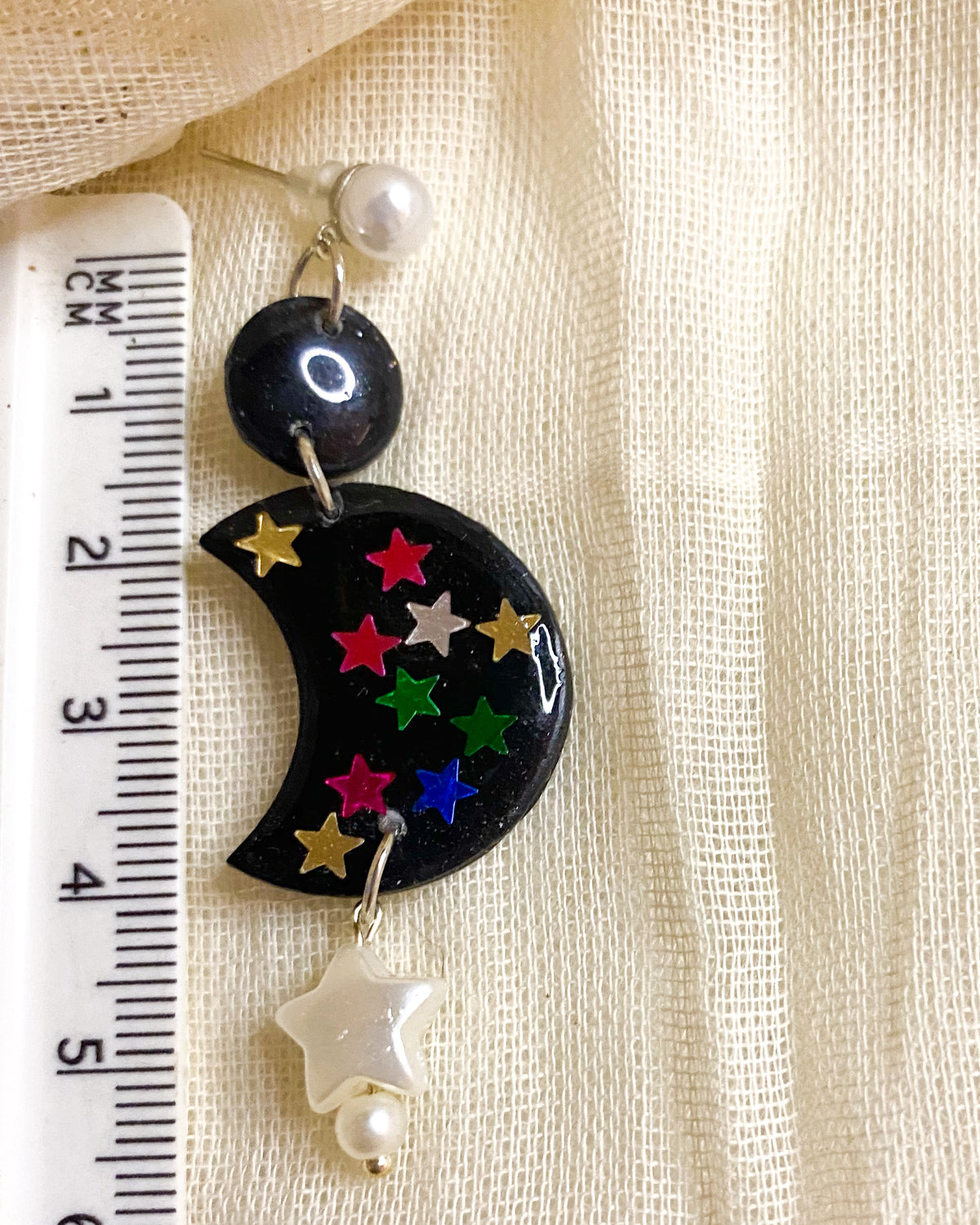 Close-up of black glitter crescent moon clay earring with colourful stars, pearl-style stud and white star drop beside a ruler