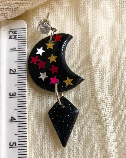 Close-up of black glitter clay earring with colourful stars, crescent moon and diamond-shaped drop beside a ruler