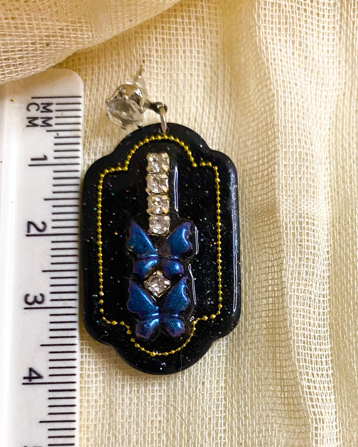 Close-up of black glitter clay dangle earring with blue butterflies, clear crystal-style gems and gold-tone beaded outline beside a ruler