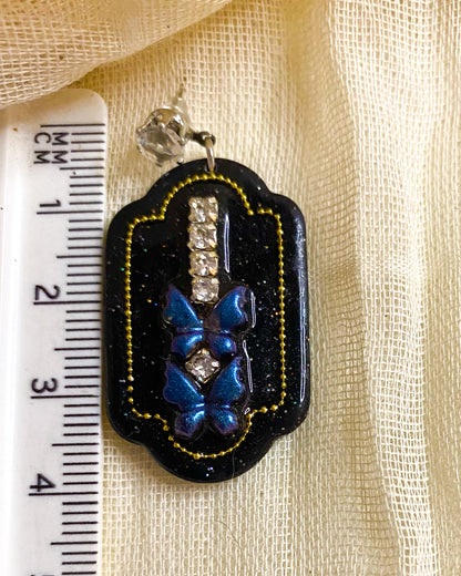 Close-up of black glitter clay dangle earring with blue butterflies, clear crystal-style gems and gold-tone beaded outline beside a ruler