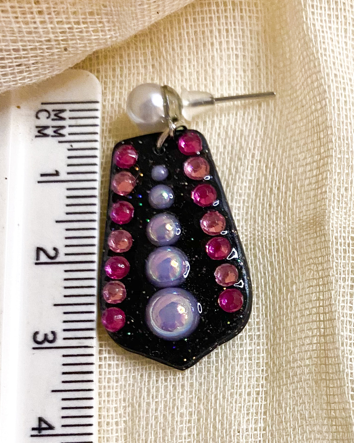 Close-up of black glitter geometric clay earring with pink gems and lilac-toned beads beside a ruler