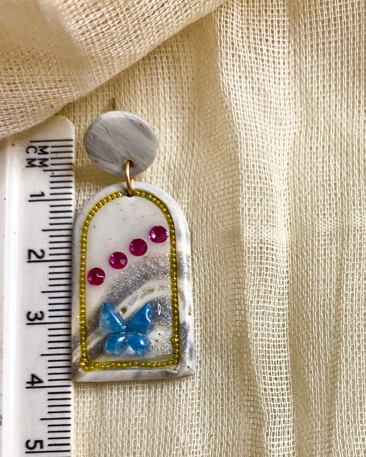 Pair of white clay arch earrings with blue butterfly details and pink gems displayed in a white circular stand