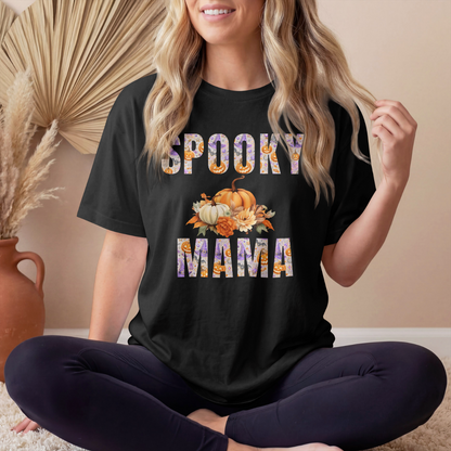 A woman sitting cross-legged indoors wearing a black graphic T-shirt with the words “SPOOKY MAMA” in a floral pattern and a pumpkin and fall leaves design in the center. The background shows a beige wall, a decorative dried palm leaf, and a terracotta vase.