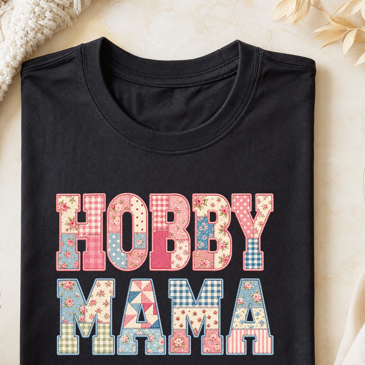 A folded black T-shirt with a colorful patchwork-style 'HOBBY MAMA' graphic on the front, styled on a light beige surface with a knit blanket and dried flowers in the corners.