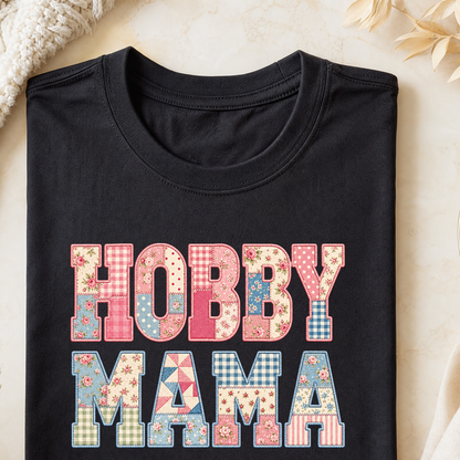 A folded black T-shirt with a colorful patchwork-style 'HOBBY MAMA' graphic on the front, styled on a light beige surface with a knit blanket and dried flowers in the corners.