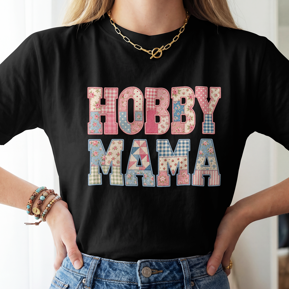 A woman wearing a black short-sleeve T-shirt with the words 'HOBBY MAMA' printed in large patchwork-style letters. The shirt is shown on a model with blonde hair, a gold chain necklace, and bracelets, paired with blue jeans.